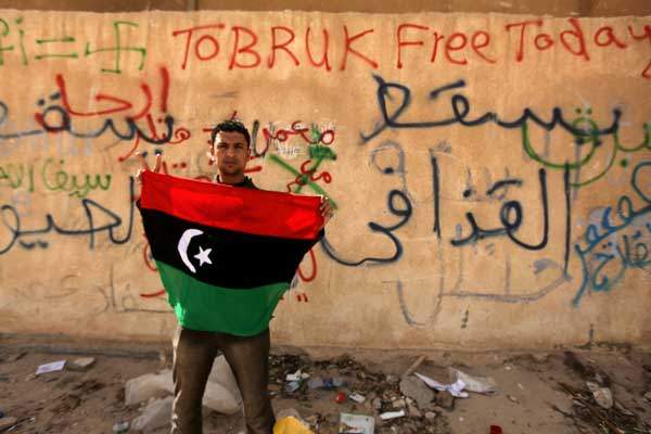 A Libyan anti-government protester holds his old national flag in front of a wall covered with graffiti against Libyan leader Moammar Gadhafi in the eastern city of Tobruk on February 24, 2011.