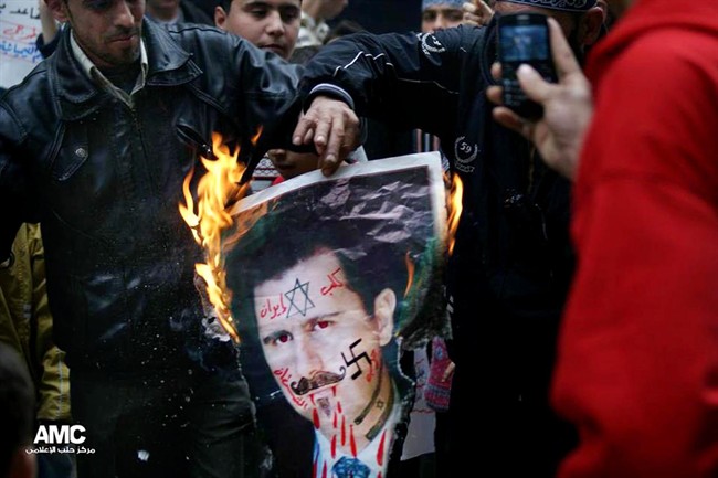 In this Monday March 25, 2013, citizen journalism image provided by Aleppo Media Center AMC which has been authenticated based on its contents and other AP reporting, anti-Syrian regime protesters burn a portrait for Syrian President Bashar Assad during a protest, in Aleppo, Syria. Syrian activists say government forces have seized control of a neighborhood in the central city of Homs that is a symbol of opposition to President Bashar Assad's regime. The Arabic on the poster reads, the dog of Iran, devil's party (Hezbollah)." (AP Photo/Aleppo Media Center, AMC).