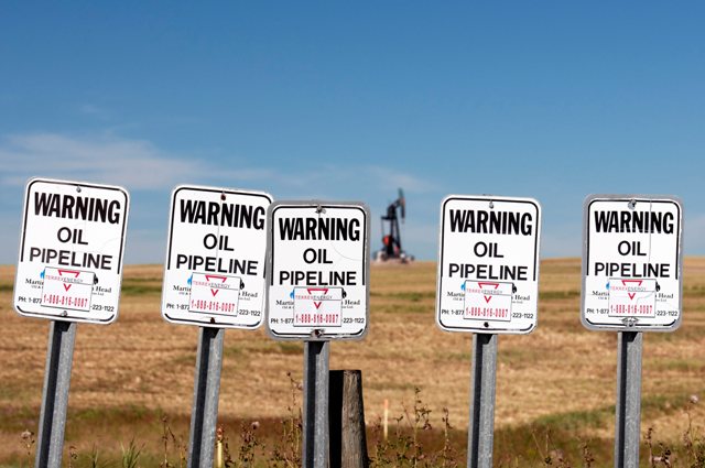Signs indicate the presence of oil pipelines near the hamlet of Carseland;Alberta. In the distance an oilfield pumpjack works pumping crude oil.