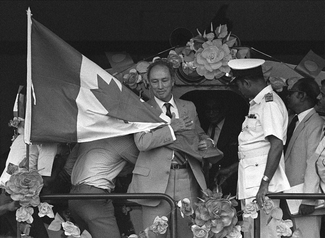 Former Canadian Prime Minister Pierre Trudeau hugs a Canadian flag that blew in his face while riding a train at a railroad re-engineering factory sponsored by CIDA in Dhaka Nov. 20, 1983.
