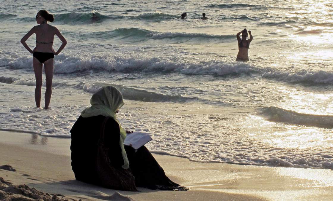 FILE - In this Thursday, Sept. 6, 2012 file photo, a woman reads the Quran on a beach as people in bikinis sunbathe and swim at sunset in Dubai, United Arab Emirates. Bikinis and briefs are no longer welcome on some sands in the United Arab Emirates. Authorities in Ras al-Khaimah, the northernmost emirate in the UAE, have posted signs on public beaches warning of possible fines for revealing swim wear such as two-piece bikinis for women and brief trunks for men.