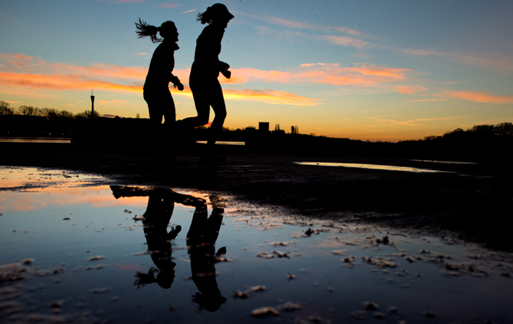 Two joggers are silhouetted against the morning sky and reflect in a puddle.