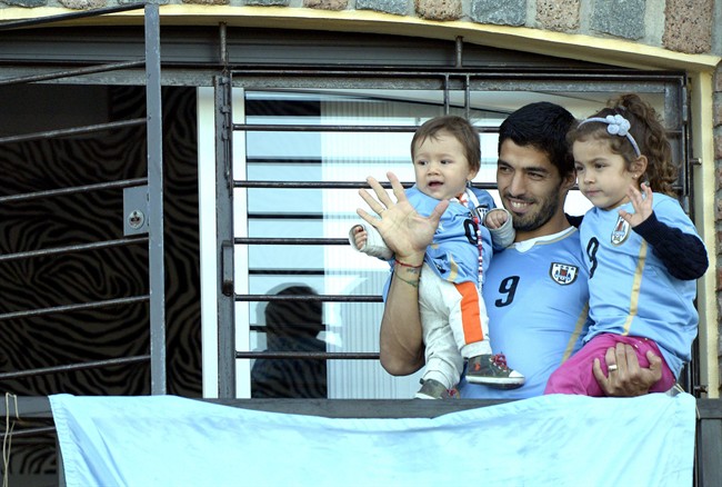 Uruguay's soccer player Luis Suarez, center, with his children Benjamin, left, and Delfina, waves to fans from his home, before the start of his team's World Cup round 16 match with Colombia, on the outskirts of Montevideo, Uruguay, Saturday, June 28, 2014. FIFA banned Suarez from all football activities for four months for biting an opponent at the World Cup, a punishment that rules him out of the rest of the tournament. (AP Photo/Matilde Campodonico)