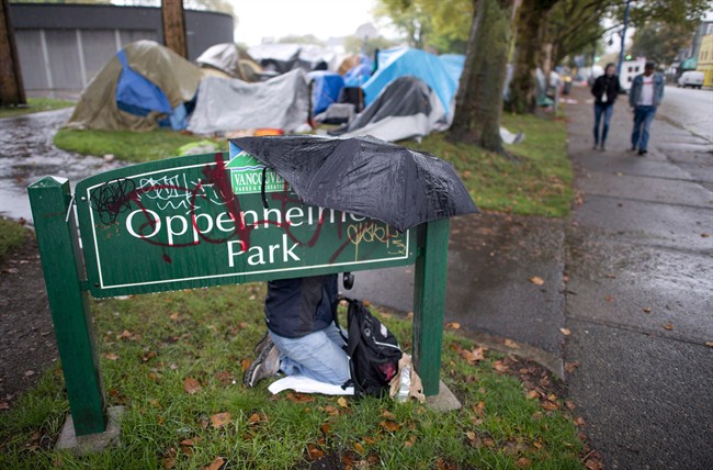 A woman takes cover from the rain behind a park sign in Oppenheimer Park in downtown Vancouver, Friday, Sept. 26, 2014. Vancouver's police force says some of its officers will be wearing video cameras during the dismantling of a homeless camp that is facing a court-ordered eviction. THE CANADIAN PRESS/Jonathan Hayward.