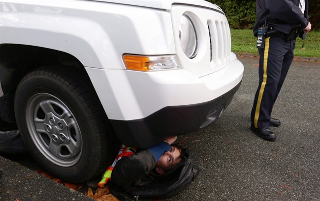 An RCMP officer stands by as a protester lies under a vehicle being used by surveyors working on behalf of Kinder Morgan in preparation for the Trans Mountain Pipeline expansion project on Burnaby Mountain in Burnaby, B.C., on Wednesday October 29, 2014. THE CANADIAN PRESS/Darryl Dyck.