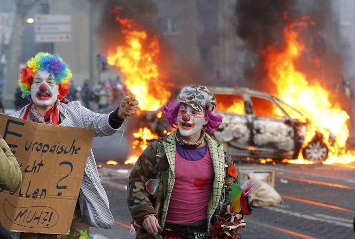 Demonstrators dressed as clowns pass by a burning police car Wednesday, March 18, 2015 in Frankfurt, Germany. 