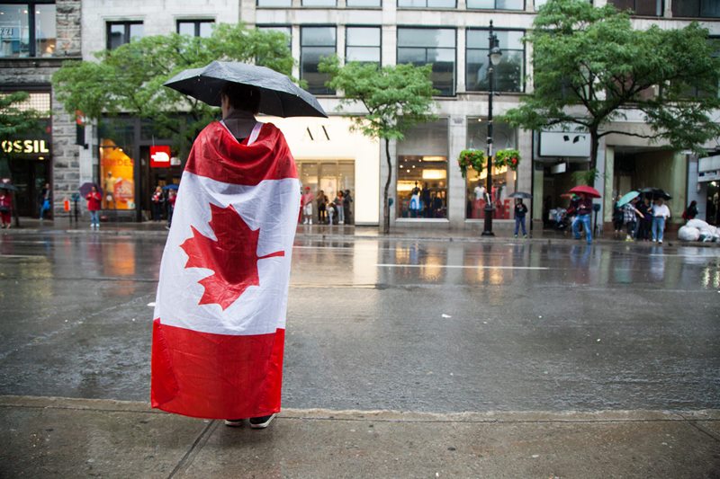 A man wrapped in his Canadian flag stands alone under an umbrella on Sainte-Catherine street.