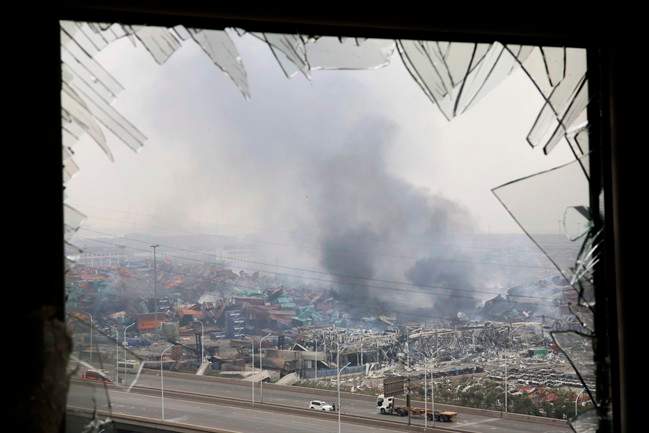 In this photo taken Friday, Aug. 14, 2015, a window shattered by the shockwaves frame the site of an explosion at a warehouse in northeastern China's Tianjin municipality.