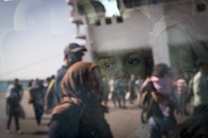 A little girl from Syria looks out of a bus as the ferry she arrived in is reflected in the bus window at the port of Piraeus, Greece, Tuesday, Aug. 25, 2015. About 2,400 Syrian refugees  stranded on Lesbos _ which they reached in small boats from nearby Turkey _ due to a dearth of ferry tickets in the high holiday season, were on the ferry. Greece has been overwhelmed this year by record numbers of migrants. (AP Photo/Petros Giannakouris).