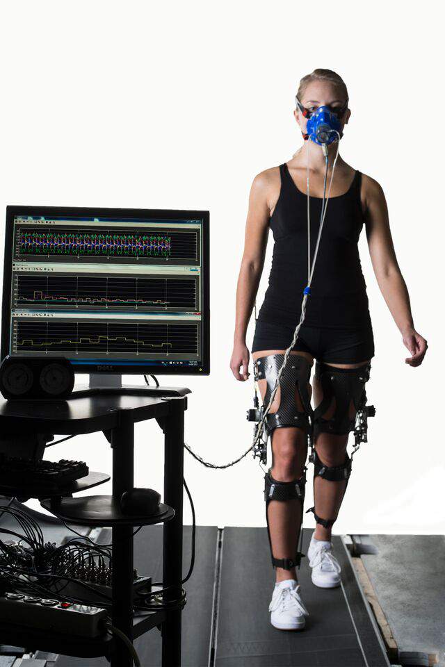 A woman is seen wearing an exoskeleton and other instrumentation measuring her energy output as she walks on a treadmill.