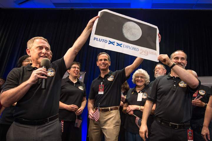 Pluto Explored. (left to right): New Horizons Principal Investigator Alan Stern of Southwest Research Institute (SwRI), Boulder, Colorado; New Horizons’ Deputy Project Scientist Leslie Young, SwRI; Johns Hopkins University Applied Physics Laboratory (APL) Director Ralph Semmel; Annette Tombaugh, daughter of Clyde Tombaugh, who discovered Pluto in 1930; and New Horizons Co-Investigator Will Grundy, Lowell Observatory, Flagstaff, Arizona hold a print of the 1991 Pluto stamp –with their suggested update – on July 14 at APL in Laurel, Maryland.