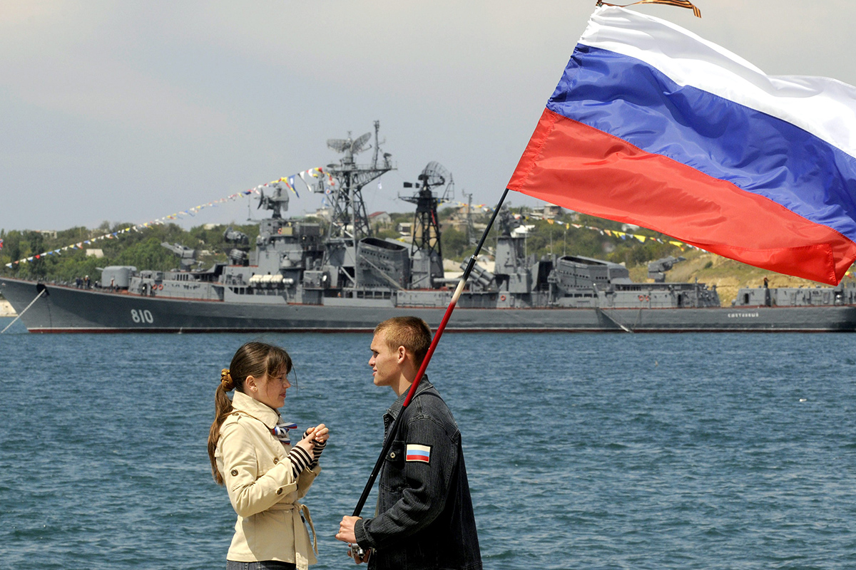 A man holding a Russian flag speaks with a girl wearing a telnyashka, a traditional navy sailor's striped vest, under her jacket during a naval parade to mark the 225th anniversary of Russian navy's Black Sea fleet at the Crimean Peninsula port of Sevastopol, Ukraine, Sunday, May 11, 2008, with the Russian Smetlivy destroyer in the background.  