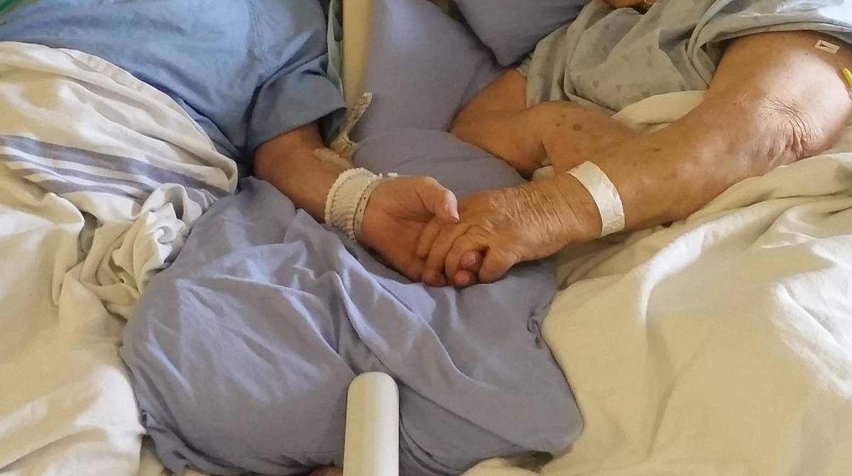 A couple hold hands in a hospital bed.
