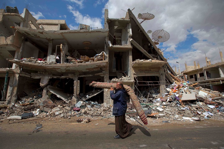 In this picture taken Thursday, April 14, 2016, a Syrian man carries a carpet as walk through a devastated part of the town of Palmyra as families load their belongings onto a bus in the central Homs province, Syria. Thousands of residents of this ancient town who fled Islamic State rule are returning briefly to check on their homes and salvage what they can.
