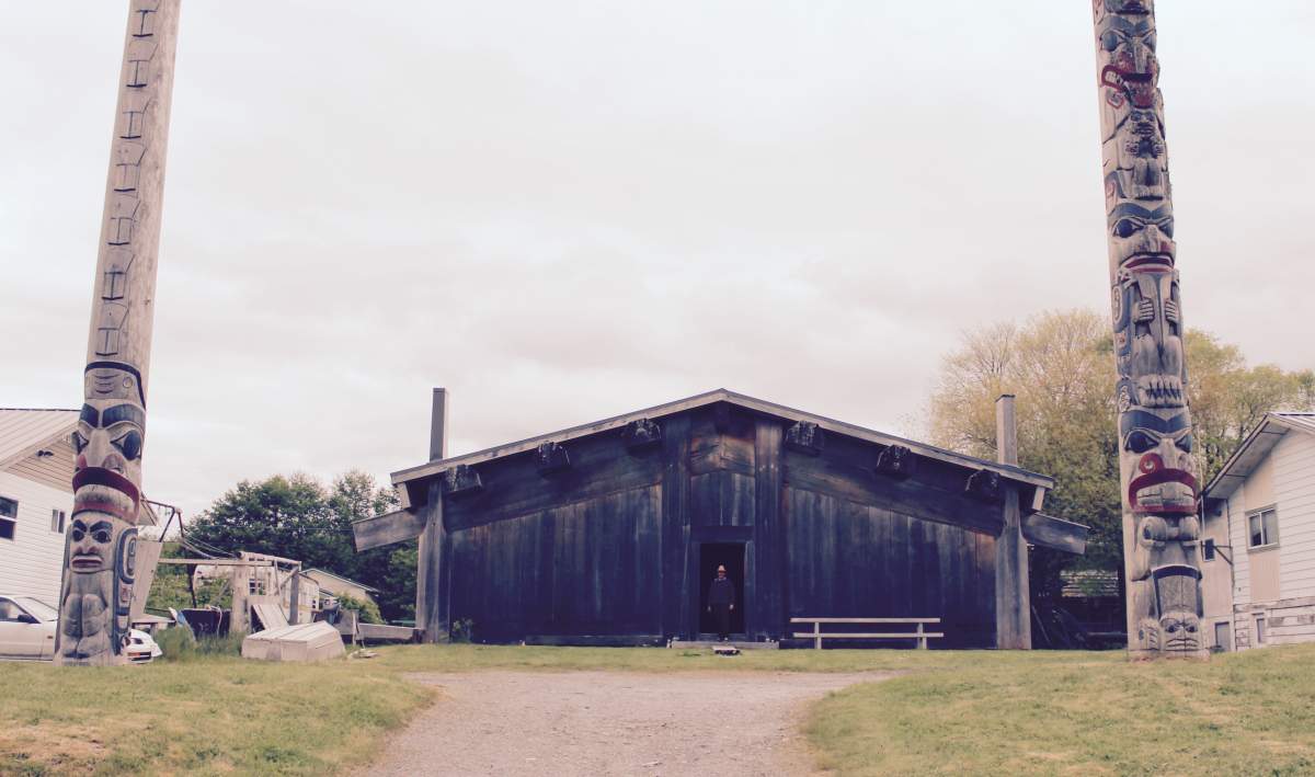Haida carver Christian White stands in front of his longhouse and the two totem poles he carved decades ago. 