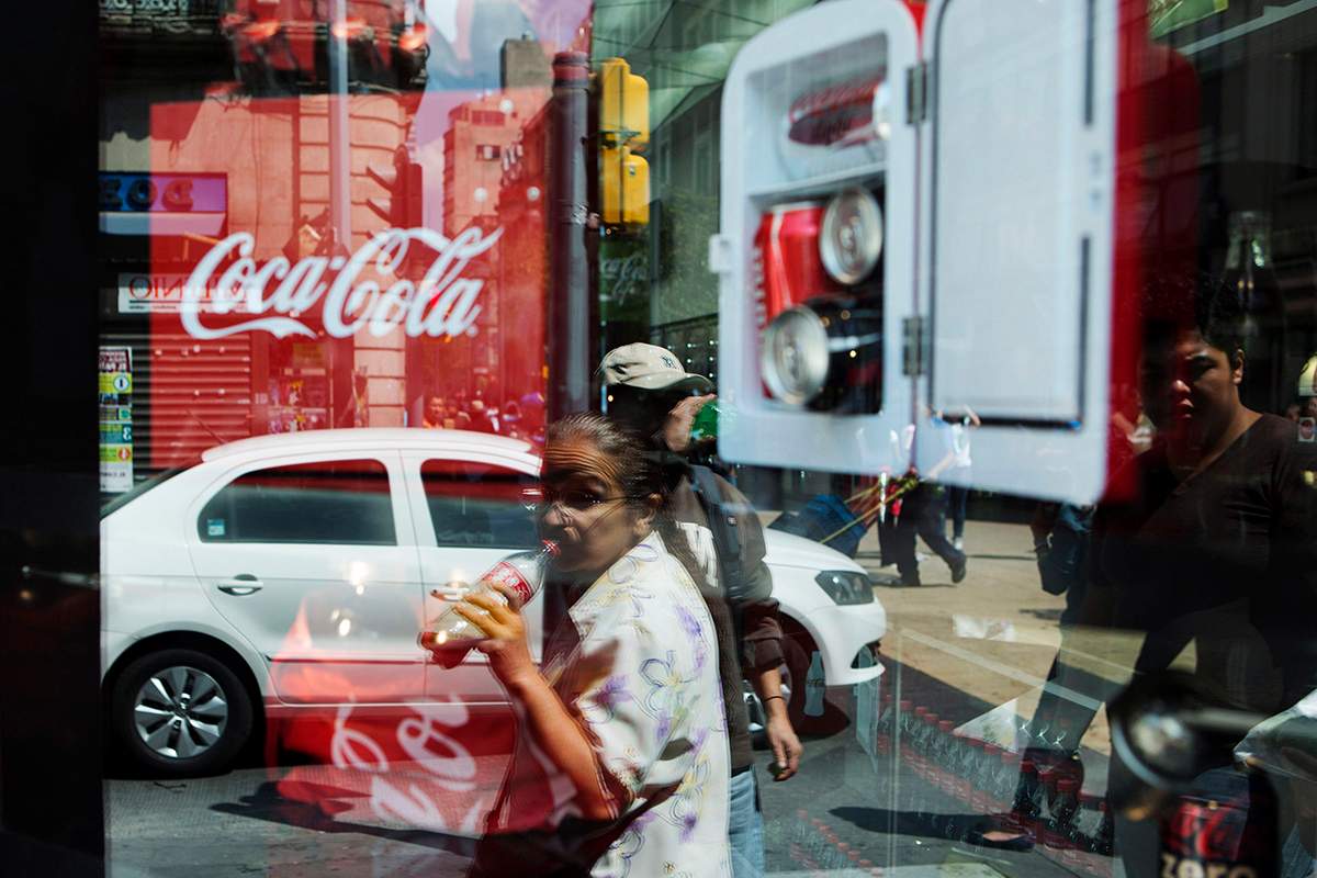 A woman is reflected in a Coca-Cola store window display as she drinks a Coke in Mexico City, Thursday, Oct. 9, 2014.