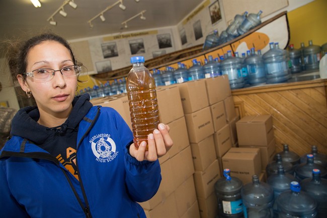 Potlotek First Nation resident Patricia Paul displays a sample of water she says came from her taps at home as she poses for a photo in front of bottles of water being supplied on demand to Potlotek residents in St. Peters, N.S., on Tuesday, October 4, 2016. 