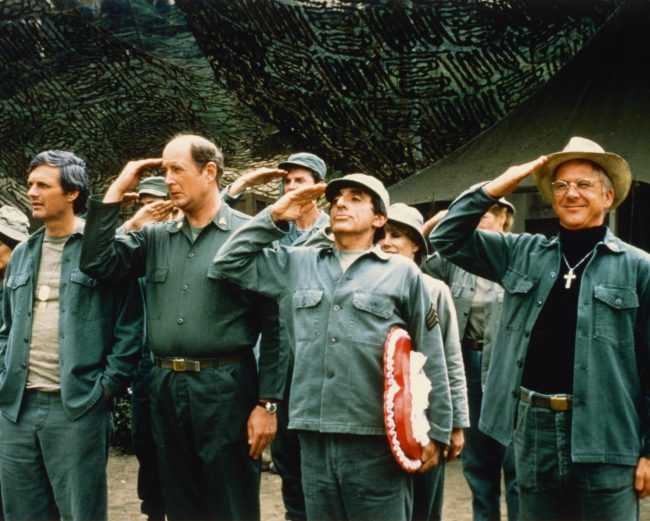 William Christopher, wearing a hat, salutes with his co-actors in a 1975 publicity image for 'M*A*S*H*'.