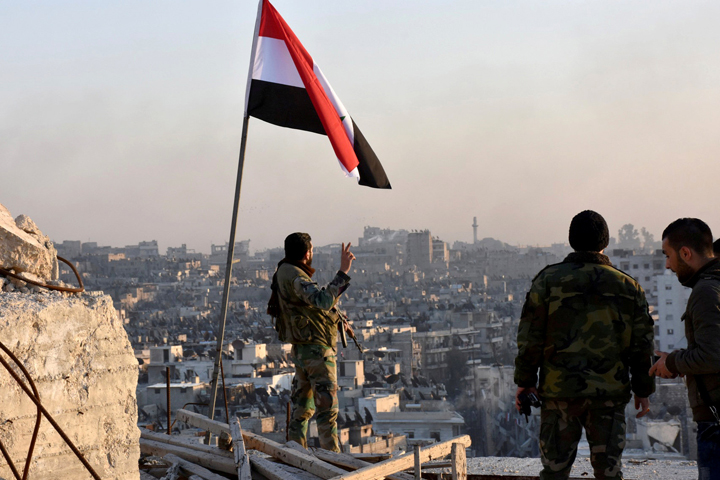 A Syrian government soldier gestures under the Syrian national flag near a general view of eastern Aleppo after they took control of al-Sakhour neigbourhood in Aleppo, Syria in this handout picture provided by SANA on November 28, 2016.