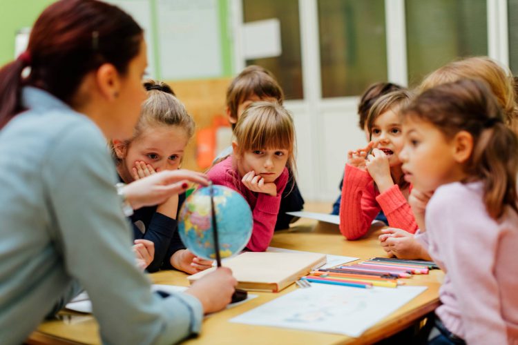 A teacher holds a globe as kindergarten students gather around her. 