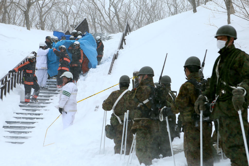 Firefighters carry a survivor they rescued from the site of an avalanche in Nasu town, Tochigi prefecture on March 27, 2017, while Self Defense Force personnel look on. 
Eight high school students were feared dead on March 27 after being engulfed by an avalanche while on a mountain-climbing outing with dozens of others, officials said. / AFP PHOTO / JIJI PRESS / JIJI PRESS / Japan OUT        (Photo credit should read JIJI PRESS/AFP/Getty Images).