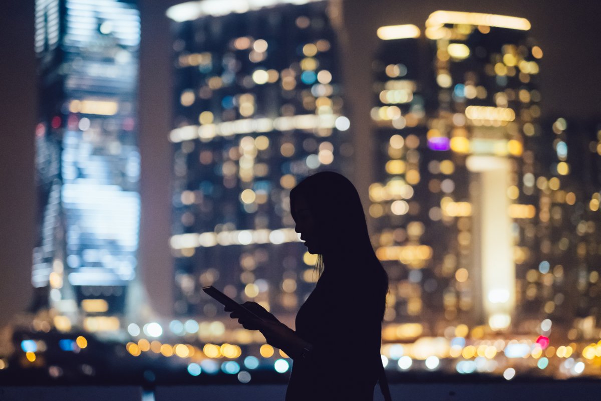 Silhouette of woman using digital tablet device in city at night, with illuminated and blurry commercial skyscrapers in the background.