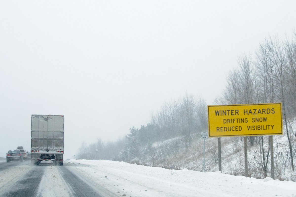 A sign warning of winter hazards - drifting snow and reduced visibility - on Highway 401 in Ontario, Canada, with the rear of a transport truck in the distance of the snow-covered roadway.