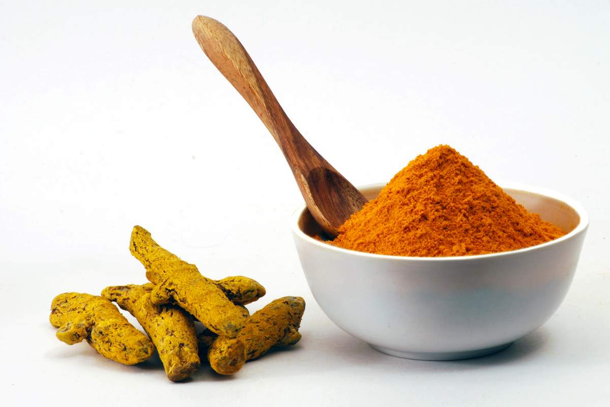 Turmeric Powder in bowl and raw turmeric with wooden spoon against white background.