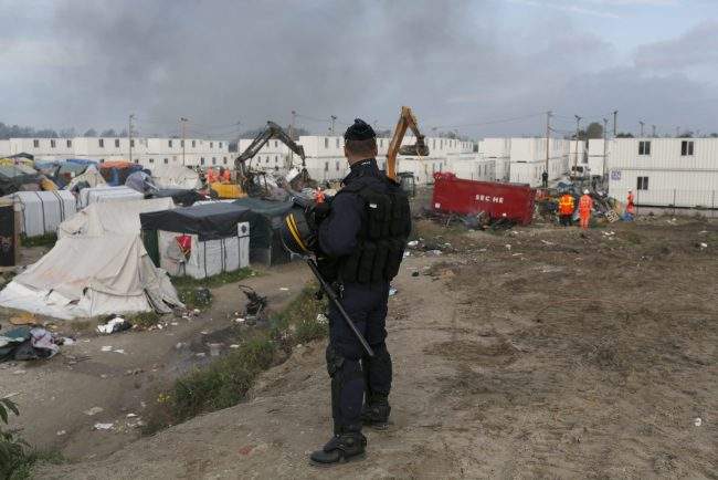 In this Oct. 26, 2016 file photo, a police officer stands guard as workers dismantle structures of the makeshift refugee camp known as the 'Jungle' in Calais, France.  