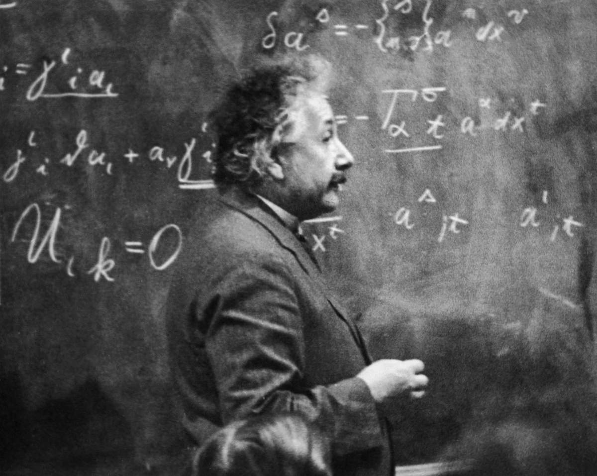 German-born physicist Albert Einstein (1879-1955) standing beside a blackboard with chalk-marked mathematical calculations written across it.