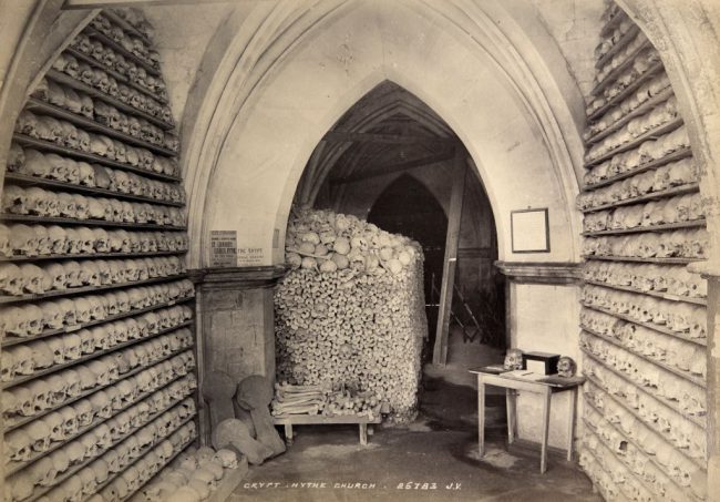 The ossuary at St. Leonard's Church in Hythe, Kent, U.K., photographed in the year 1900. The photograph shows advertisements for guides written by the vicar, and a table with what appears to be a visitors' book and a donations box.
