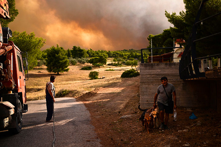 A man walks his dog past a man holding a hose as smoke billows in background during a wildfire in Kineta, near Athens, on July 23, 2018.