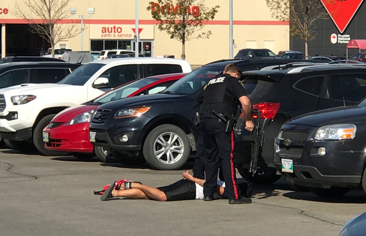 Two police officers stand over a man in the Polo Park Shopping Centre parking lot. One officer is holding a large rifle.