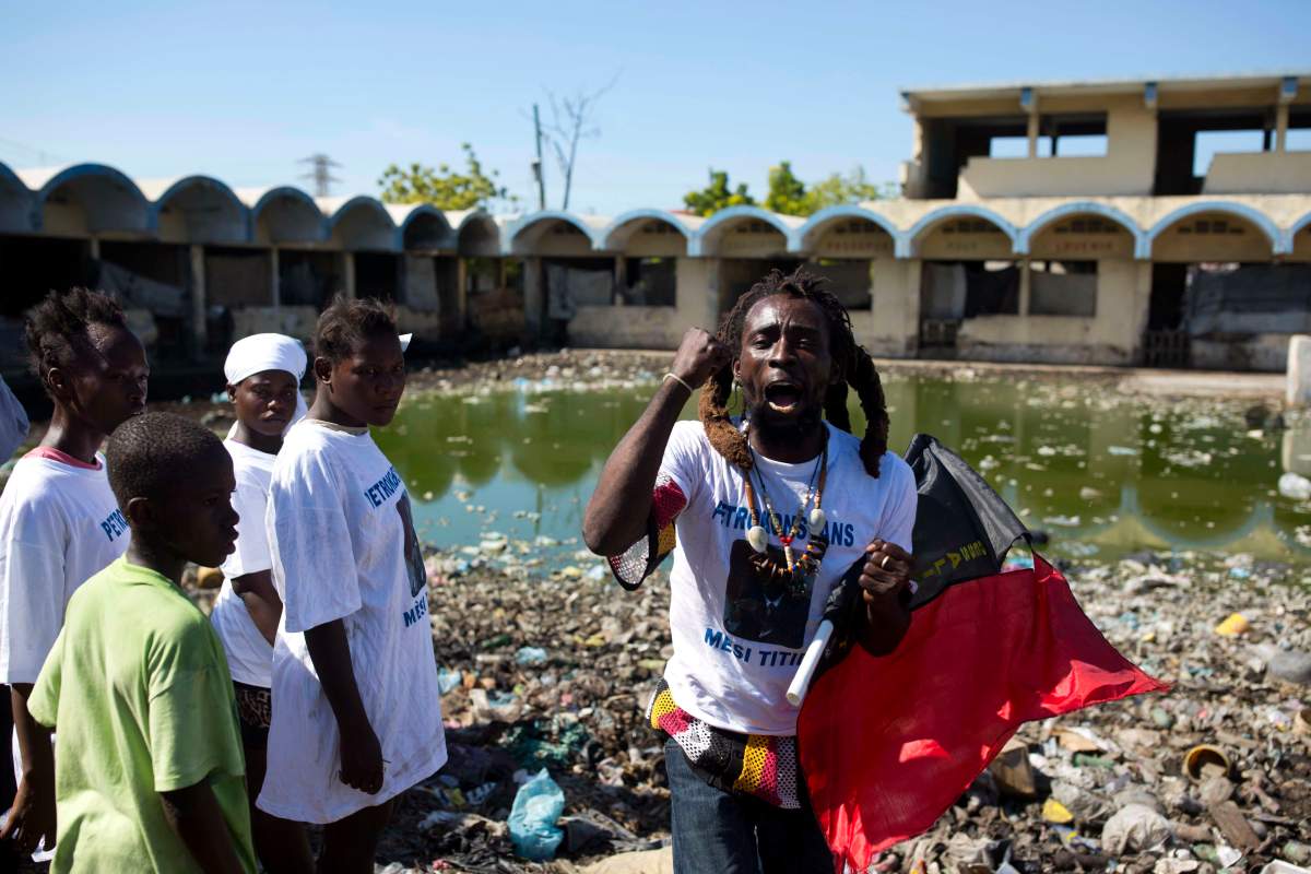 In this Dec. 16, 2018, photo, a man wearing a T-shirt showing former president Jean Bertrand Aristide and clutching a flag associated with the Pitit Dessalines political party, decries the government as people gather at a pig farm, once a school, where the victims of a massacre were fed to the animals in the La Saline slum of Port-au-Prince, Haiti. Some residents and local rights groups accuse mid-ranking officials of orchestrating the Nov. 13 attack by a rival gang to intimidate residents and prevent anti-government protests.
