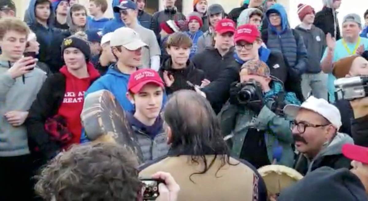 In this Friday, Jan. 18, 2019, image made from video provided by the Survival Media Agency, a teenager wearing a Make America Great Again hat, centre left, stands in front of an elderly Native American singing and playing a drum in Washington, D.C.