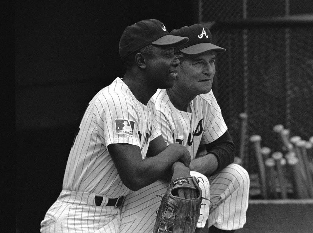 In this Sept. 30, 1969, file photo, a Major League Baseball 100th anniversary patch is shown on the uniform of Atlanta Braves team captain Hank Aaron, left, as he watches from the dugout with manager Luman Harris, during a game against the San Diego Padres, in Atlanta. (AP Photo/Joe Holloway Jr., File)