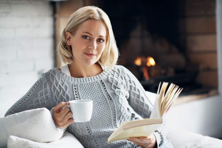 A young woman enjoying her novel with a hot drink while sitting inside near a fireplace.