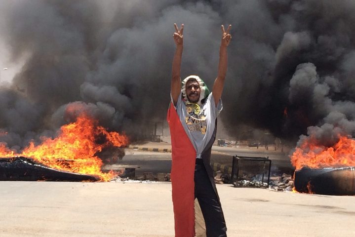 In this June 3, 2019 file photo, a protester flashes the victory sign in front of burning tires and debris on road 60, near Khartoum’s army headquarters, in Khartoum, Sudan.