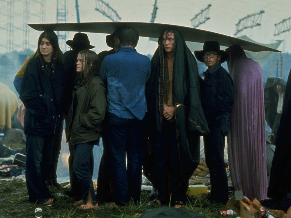 A sodden group, seeking shelter from rain, using a sheet of plywood as a makeshift umbrella, at the iconic Woodstock 1969 music festival.