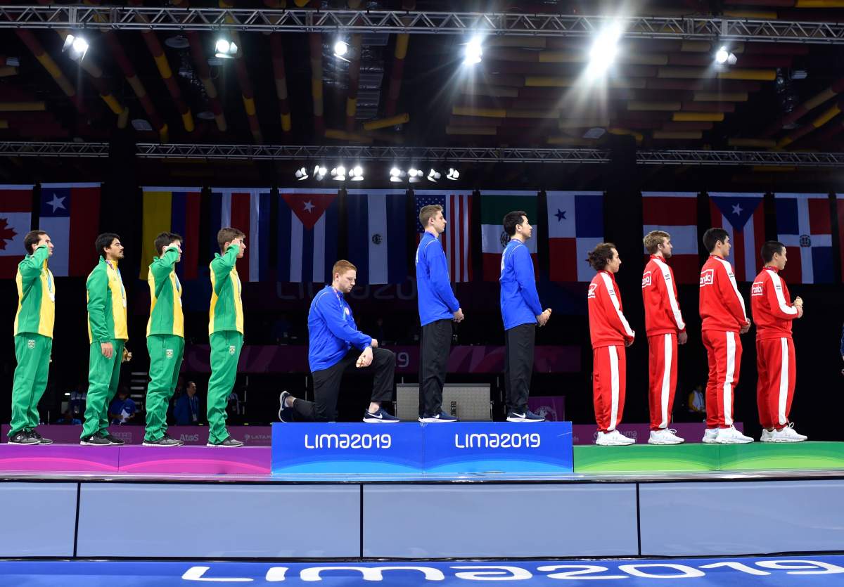 In this Friday, Aug. 9, 2019 photo, released by Lima 2019 News Services, Race Imboden of the United States takes a knee, as teammates Mick Itkin and Gerek Meinhardt stand on the podium after winning the gold medal in team's foil, at the Pan American Games in Lima, Peru. 