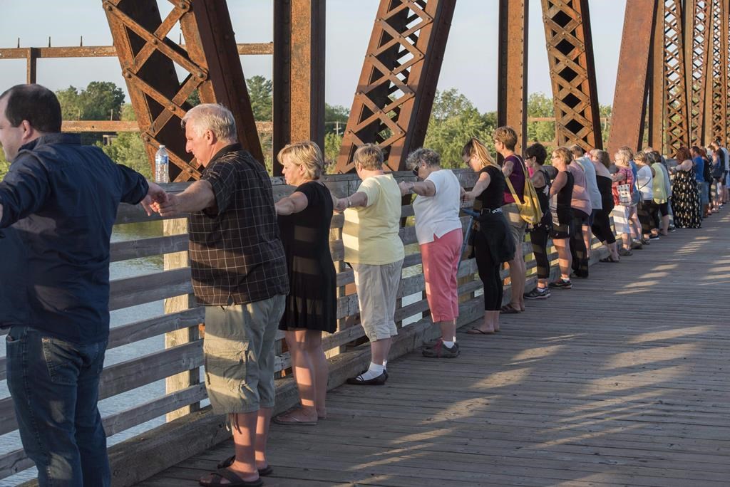 Mourners join hands as thousands of people hold hands to remember the Aug. 10, 2018, shooting victims by joining hands on the Bill Thorpe Walking Bridge in downtown Fredericton on Monday, August 13, 2018. Hands and Hearts Across the City, an event held just days after the shootings, will be held again Saturday. THE CANADIAN PRESS/Stephen MacGillivray.
