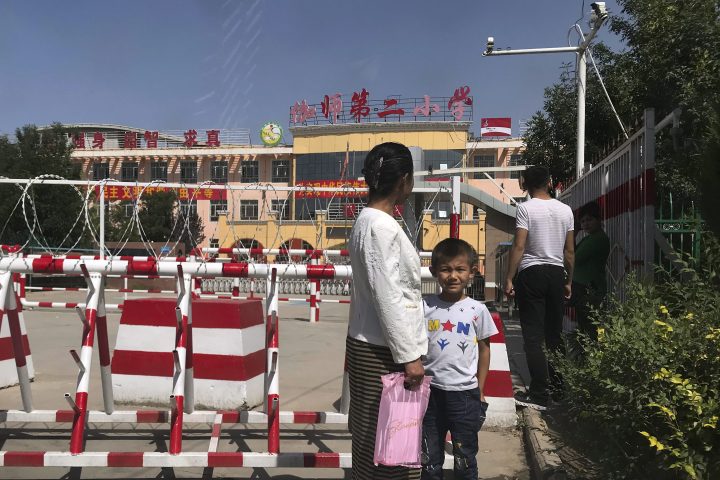 In this Aug. 31, 2018, file photo, a child and a woman wait outside a school entrance mounted with surveillance cameras and barricades with multiple layers of barbed wire in Peyzawat, western China's Xinjiang region.  