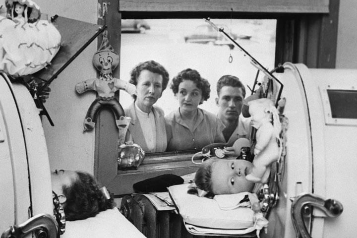 Two little girls lie in iron lung machines while being treated for polio in 1952. Their family looks in from the window outside.