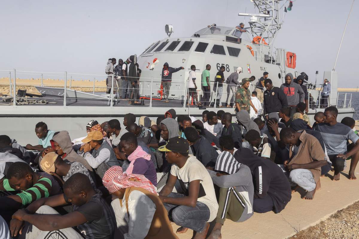 In this Oct. 1, 2019 photo, rescued migrants are seated next to a coast guard boat in the city of Khoms, around 120 kilometers east of Tripoli, Libya.
