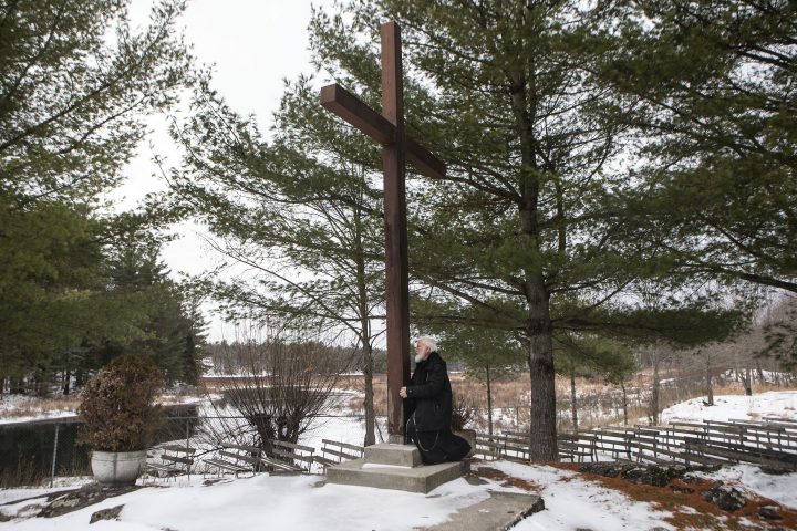 Father Claudio Piccinini, a Catholic priest and founder of “Teopoli, the City of God,” a religious place of worship, in Gravenhurst, Ont., kneels at the base of a large cross on the grounds, on Friday, December 13, 2019.