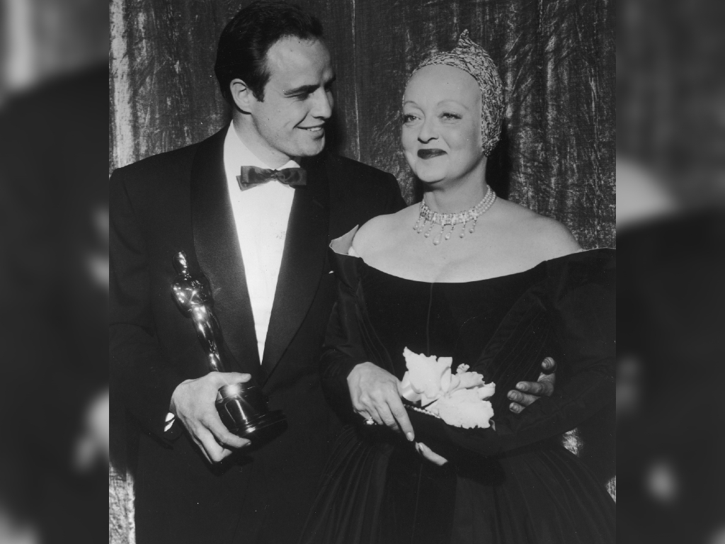 American actors Marlon Brando and Bette Davis smile together backstage at the Academy Awards. Brando holds the Oscar statuette he won for Best Actor for ‘On the Waterfront.’