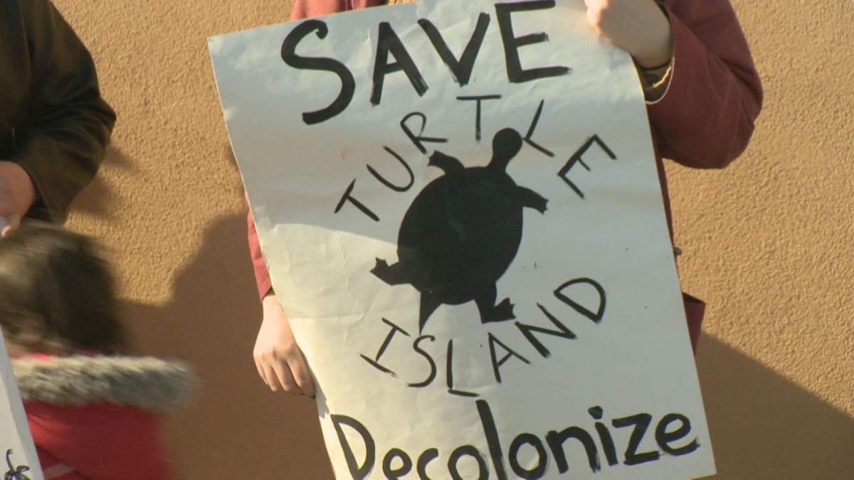 A protester stands in front of Andrew Scheer’s constituency office on Feb. 22 while holding a sign that reads “Save Turtle Island, Decolonize”