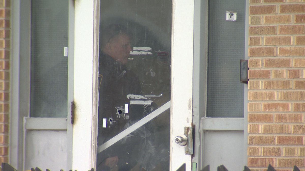A police officer is seen behind the front door of a Winnipeg apartment building, which appears to have bullet holes.