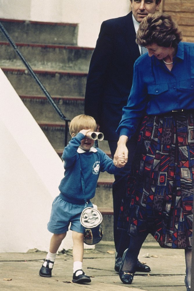 In this Sept. 16, 1987 file photo, Britain’s Prince Harry looks through mock binoculars while holding the hand of kindergarten manager Jane Mynors and carrying a Thomas the Tank Engine bag on his way out from his first day at a kindergarten in Notting Hill, West London. (AP Photo/Martin Cleaver, File)