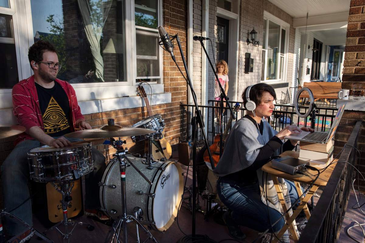 As neighbor Carys Williams, 7, peeks out of her house, drummer Andrew Northcutt, left, waits as singer Elena Lacayo, works on technical issues for their band, Elena and Los Fulanos, as they tried to live stream a concert from the porch of their house, Sunday April 5, 2020, in Washington. The band, who normally make their income from live concerts and touring, is trying to make some income under coronavirus restrictions by live streaming concerts and asking for contributions from their viewers. 